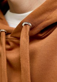 Närbild av en brun hoodie med en strukturerad tyg, metallögla och vävd snörning. Designen inkluderar ett vitt innerlager som är synligt vid halsen.