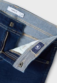 Džinsu bikses ar tumši zilas krāsas galveno audumu, gaiši zilas krāsas jostu, ar rāvējslēdzēju un pogu detaļu. Iekšpusē ir mīksts, gaiši zils odere.