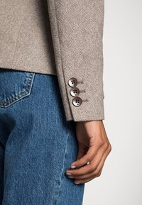 Blazer beige a texture con un fit su misura, caratterizzato da tre bottoni marroni sui polsini, abbinato a jeans in denim blu.