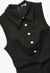 Musta hihaton mekko, jossa on kauluskohouma, viisi kiiltävää napin aksenttia ja kangasvyö vyötäröllä. Silkkinen tekstuuri.