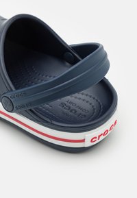 Navy blue Crocs clog with adjustable heel strap, white sole with red stripe, and red Crocs logo on heel tab.