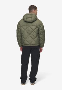 Giacca imbottita verde oliva con cappuccio, caratterizzata da una texture a rombi, polsini elastici e vestibilità rilassata, abbinata a pantaloni cargo neri.