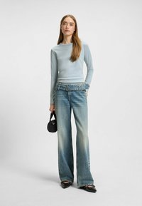 Helles blaues Langarmoberteil, ausgebleichte blaue weite Jeans mit einem gerafften Denim-Taillenbund, schwarze Handtasche und spitze schwarze Schuhe.