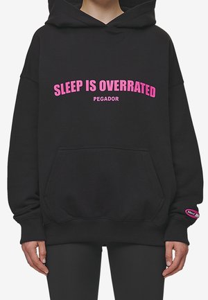 Personne portant un sweat à capuche noir avec un texte rose « LE SOMMEIL EST SURÉVALUÉ » et « PEGADOR » sur le devant ainsi qu’un petit patch sur la manche gauche.