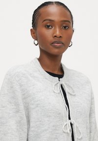 Ανοιχτό γκρι ζακέτα με υφή πλέξης, που διαθέτει στρογγυλό λαιμόκοψη και κλείσιμο με κορδόνι. Εμφανίζει ένα διακριτικό, απαλό φινίρισμα υφάσματος.