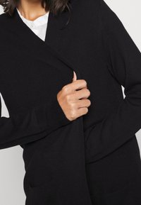 Svart stickad kofta med långa ärmar, djup V-hals och framfickor. Tyget ser mjukt och texturerat ut.