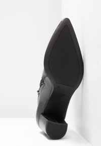Chaussure à talon haut en cuir noir avec bout pointu, semelle texturée et talon bloc proéminent. Présente des coutures décoratives. Marque visible sur la semelle.