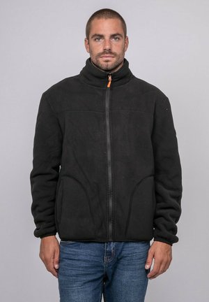 Homme aux cheveux courts debout face à l'objectif, portant une veste polaire noire zippée et un jean bleu sur un fond uni gris clair.