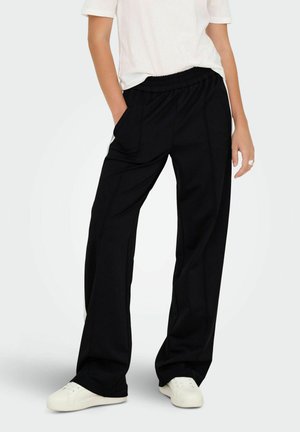 Pantalons larges noirs en tissu doux avec une taille élastique à cordon, poches latérales et coupe décontractée, associés à des baskets blanches.