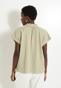 Top de manga corta en un verde oliva claro con un tejido texturizado, cuello con volantes y corte holgado, que se lleva con pantalones blancos.
