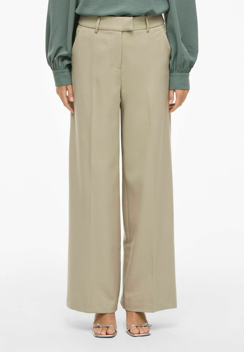 Pantalon large de couleur beige clair, avec une texture lisse, doté d'une ceinture et de poches avant, associé à un haut vert ample.