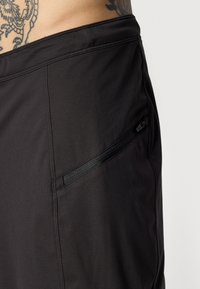 Close-up van zwarte broek met een diagonale ritszak en een gedeeltelijk zicht op een getatoeëerde torso boven de tailleband.