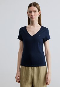 Μπλε σκούρο T-shirt με V λαιμόκοψη, κατασκευασμένο από μαλακό βαμβάκι με κοντά μανίκια, διακοσμημένο με ένα μικρό λογότυπο. Συνδυάζεται με ανοιχτόχρωμο λινό παντελόνι.