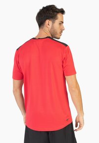 Camiseta deportiva roja de manga corta, con tejido texturizado y detalles en negro en los hombros. El diseño presenta un dobladillo redondeado y un ligero estiramiento.
