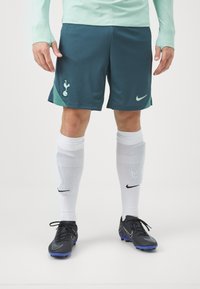 Turkoosi urheilusortsit, joissa on Tottenhamin logo ja vihreät yksityiskohdat, yhdistettyinä valkoisiin sukkiin, joissa on musta Nike-logo, ja mustiin nappulakenkiin.