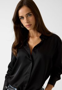 Schwarz satiniertes Hemd mit Button-Down-Kragen, langen Ärmeln und lockerer Passform. Glatte Textur und subtile Schattierung, die im Stoff sichtbar sind.