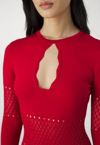 Maglietta rossa a maniche lunghe con scollo a chiave, texture a costine e motivo a diamante aperto su maniche e corpo.