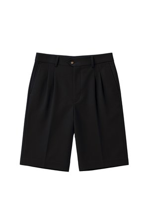 Mężczyzna ubrany w czarne szerokie szorty, białe skarpetki z napisem i czarno-białe mokasyny, stoi z rękami w kieszeniach.