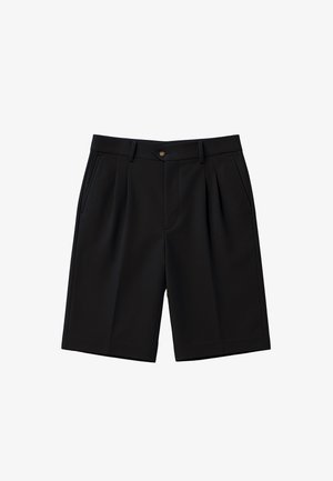 Man draagt zwarte wijd uitlopende shorts, witte sokken met tekst en zwart-witte loafers, staand met handen in de zakken.