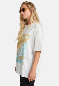 White oversized t-shirt featuring a colorful graphic of a sunset and ocean waves. Worn with brown sunglasses, showcasing a relaxed fit.