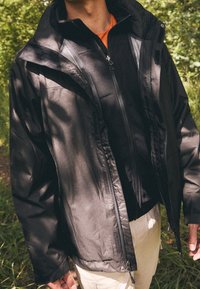 Schwarze, wetterfeste Jacke aus glattem Stoff mit Reißverschluss vorne, verstellbaren Ärmelbündchen, geschichtetem Design und weicher Innenauskleidung.