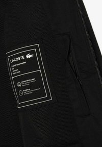 Melnā sporta jaka ar acu struktūru, ūdensizturīgu audumu, kabatu ar slēdzējiem un baltu etiķeti, kurā ir norādītas kopšanas instrukcijas un īpašības.