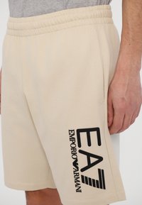 Béžové bavlněné šortky s elastickým pasem, na boku s velkým černým potiskem loga. Hladká textura s prošitým lemem.