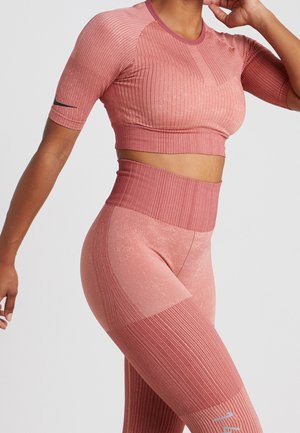 Kobieta w dopasowanym różowym prążkowanym bezszwowym crop topie i legginsach z wysokim stanem, pozująca z jedną ręką uniesioną blisko głowy.