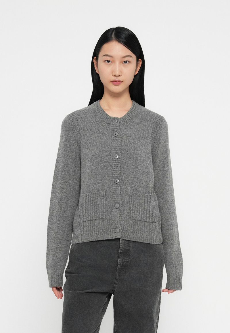 Šedý pletený cardigan s kulatým výstřihem, který má zapínání na knoflík a dvě přední kapsy s žebrovaným vzorem. Měkká textura, přiléhavý střih, dlouhé rukávy.