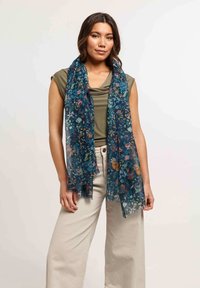 Sciarpa floreale in tessuto trasparente blu scuro, con fiori multicolore e un leggero bordo sfrangiato, indossata sopra un top verde e pantaloni beige.