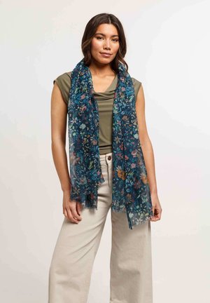 Foulard floral en tissu léger bleu foncé, présentant des fleurs multicolores et un léger bord effiloché, porté sur un haut vert et un pantalon beige.