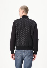 Svart bomberjacka med en texturerad bakpanel med geometriskt mönster, ribbade muddar och en helt svart front. Tillsammans med blåjeans.
