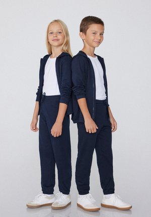 Marine athleisure set gemaakt van zachte stof, bestaande uit een ritsjack en joggingbroek met trekkoord, gecombineerd met witte en beige sneakers.