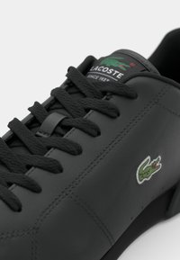 Sneaker en cuir noir avec des lacets ronds, des accents cousus et un logo crocodile brodé vert sur le côté extérieur.