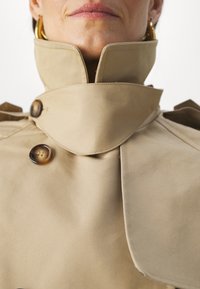Gros plan d'une personne portant un trench-coat beige avec le col relevé et des boutons en écaille, montrant les lèvres et les boucles d'oreilles.