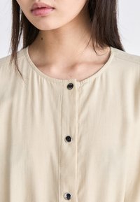 Chemise beige à boutons avec un col rond, texture lisse et boutons noirs, mettant en avant un design simple et un style minimaliste.