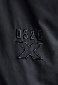 Tejido negro con el texto bordado "032C" y un diseño de "X". Textura suave, estilo minimalista, sin colores ni patrones adicionales.