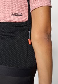 Svart og rosa treningsgenser med nettingtekstur; har Polartec-merke, sidelomme med oransje dragbånd, og tettsittende design.
