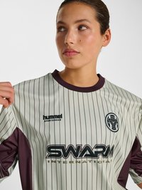 Młoda osoba nosząca beżową, wąsko prążkowaną koszulkę sportową z czarnymi logo, patrząca na bok i trzymająca kołnierz jedną ręką.