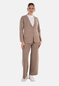 Completo sartorial beige composto da un blazer aderente con due bottoni e pantaloni a gamba larga, abbinato a un top bianco e sneakers bianche.