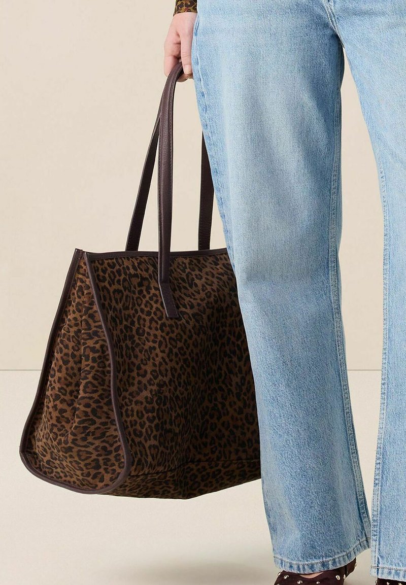 Sac fourre-tout en imprimé léopard marron avec des poignées marron foncé, présentant une forme structurée et une texture lisse. Porté avec des jeans bleu clair.