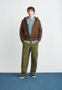 Καφέ φούτερ με φερμουάρ πάνω από ανοιχτό τζιν πουκάμισο, συνδυασμένο με χαλαρό πράσινο παντελόνι cargo και μαύρα αθλητικά παπούτσια με λευκές σόλες, στέκεται σε μπλε χαλί.