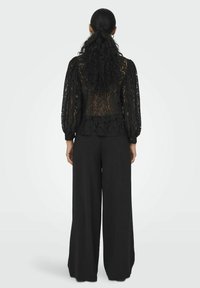 Zwarte kanten blouse met een doorschijnende rug en volumineuze lange mouwen, gecombineerd met zwarte wijde broek. Glad materiaal, elegante uitstraling.