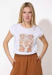 Weißes Kurzarm-T-Shirt mit einem zentralen grafischen Design in Beige- und Rosttönen mit Pflanzenmotiven. Kombiniert mit einem braunen, strukturierten Rock.