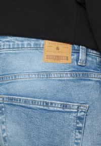 Jasnoniebieskie dżinsy z denimu z efektem przetarcia oraz brązowa skórzana metka z napisem "ONLY & SONS" przyszyta z tyłu na pasku w talii.