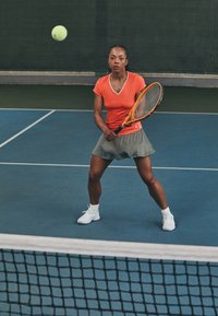 Jogadora de ténis com uma camisola de mangas curtas laranja e saia cinza segura uma raquete. Bola de ténis no ar, em um campo coberto com chão azul e rede.