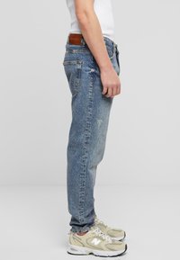 Jeanși din denim de un albastru deschis, cu o croială relaxată, având o etichetă din piele maro pe spate și o ușoară decolorare, asortați cu adidași bej.