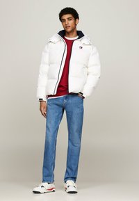 Giacca a vento bianca con colletto blu navy, maglione rosso sotto, jeans blu e sneakers bianche. Presenta cerniera e strisce decorative.