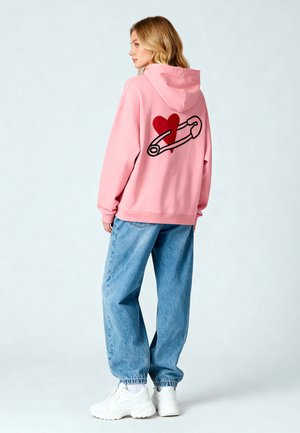 Felpa rosa con un grande cuore rosso e una spilla di sicurezza ricamati sul retro. Abbinata a jeans blu larghi e scarpe da ginnastica bianche.