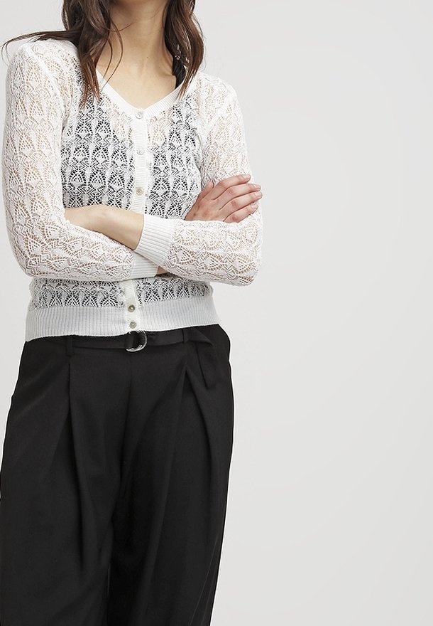 Femme les bras croisés portant un cardigan blanc en dentelle boutonné sur un haut à motifs et un pantalon plissé noir taille haute.